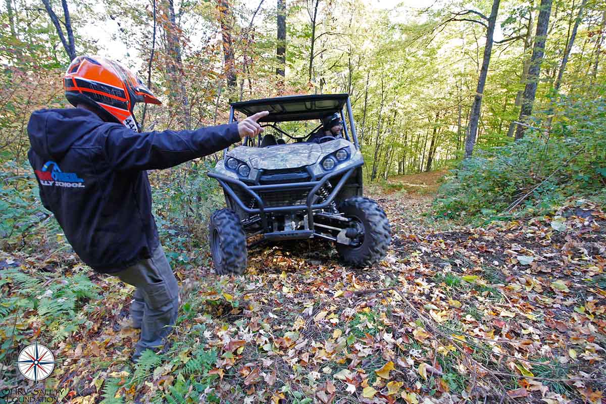 Person with a helmet and jacket pointing at an ATV in a forest
